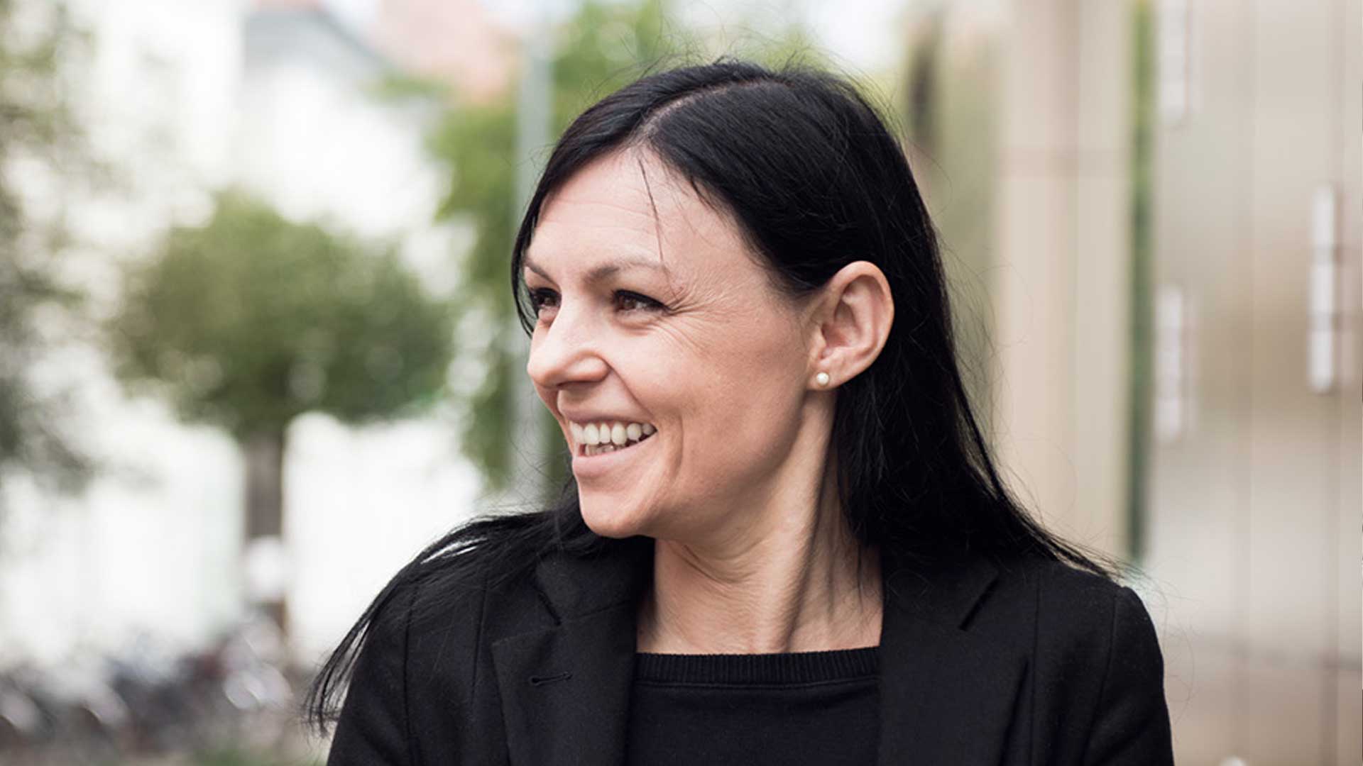 Eine Frau mit dunklen Haaren lächelt und trägt eine schwarze Jacke und ein schwarzes Hemd.